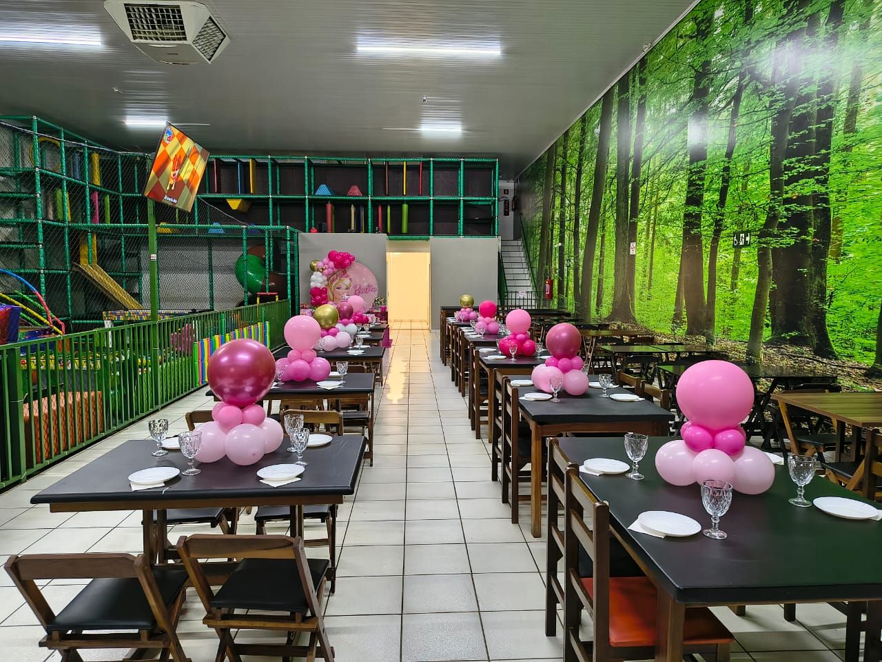 Salão do Espaço Pais & Filhos montado para festa infantil, com mesas postas e balões cor-de-rosa nos centros, o playground coberto à esquerda e a parede de floresta à direita