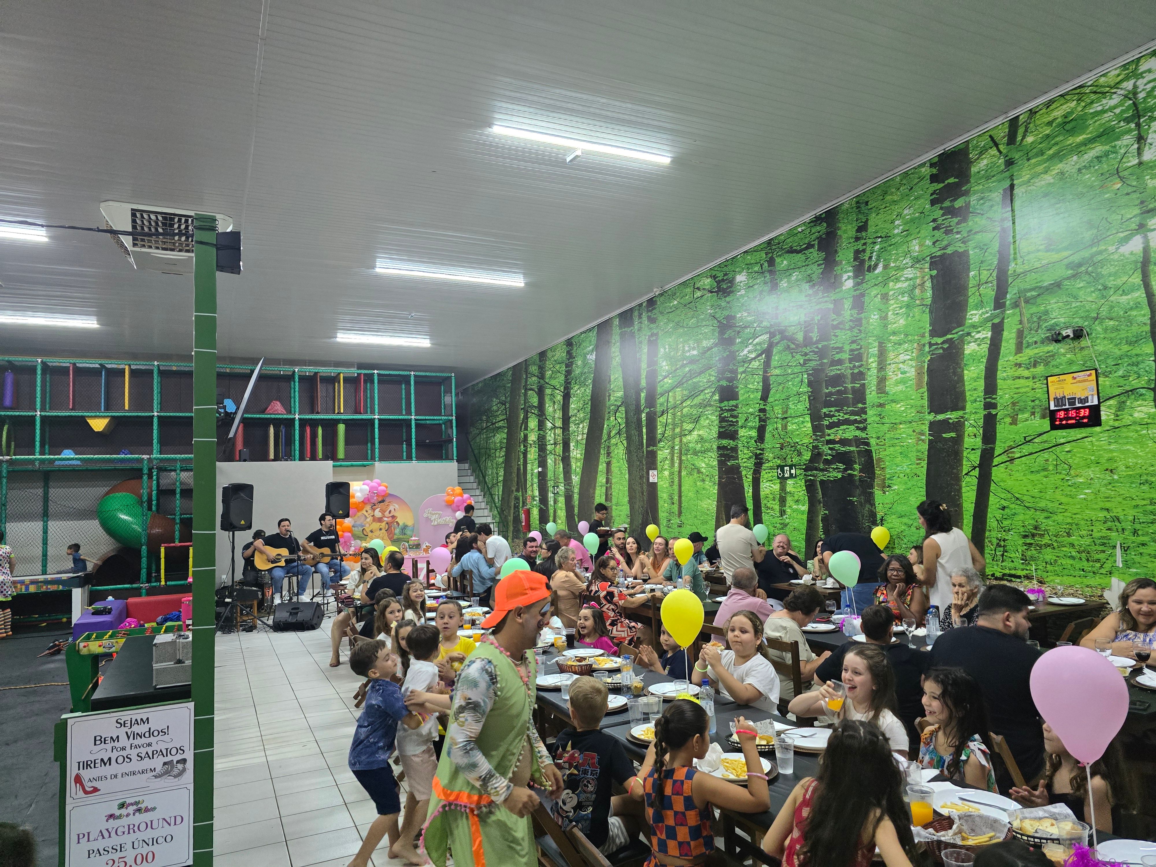 Festa lotada à noite no salão, com banda tocando ao vivo, animador dançando com as crianças e famílias às mesas