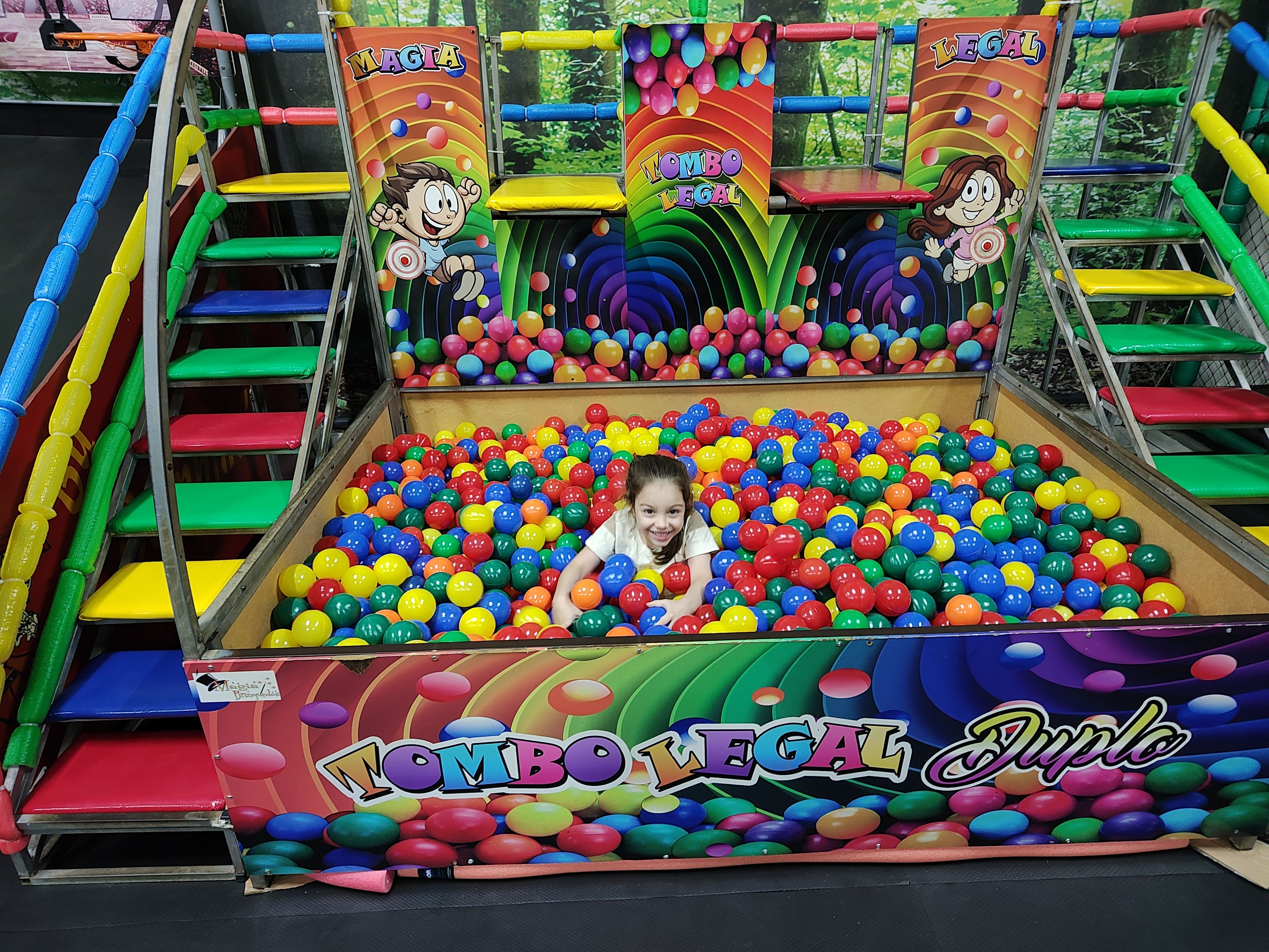 Menina brincando na piscina de bolinhas do playground coberto, com escadas e escorregadores ao redor