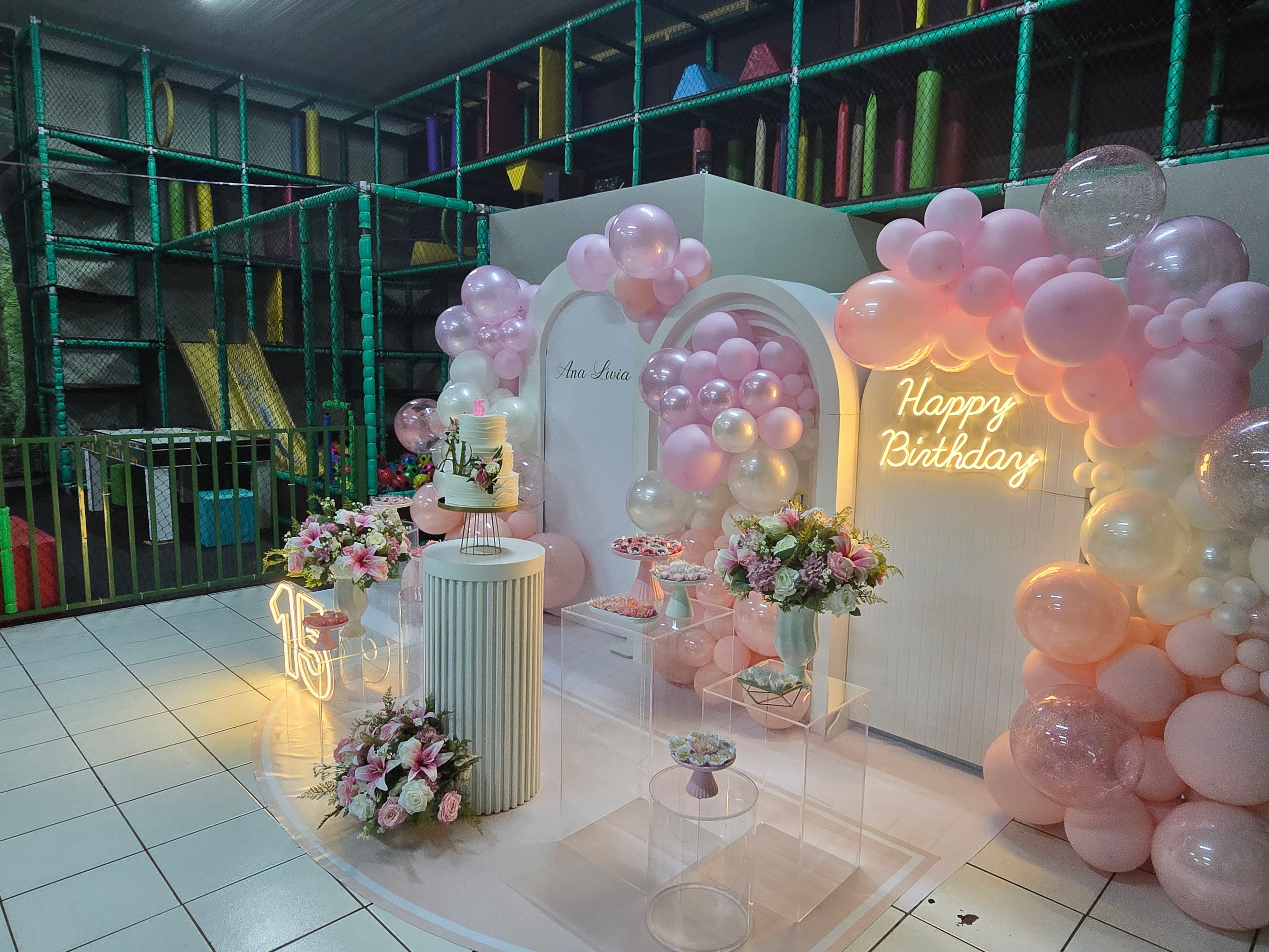 Festa de 15 anos com arco de balões cor-de-rosa, letreiro de neon, bolo e mesa de doces, com o playground ao lado