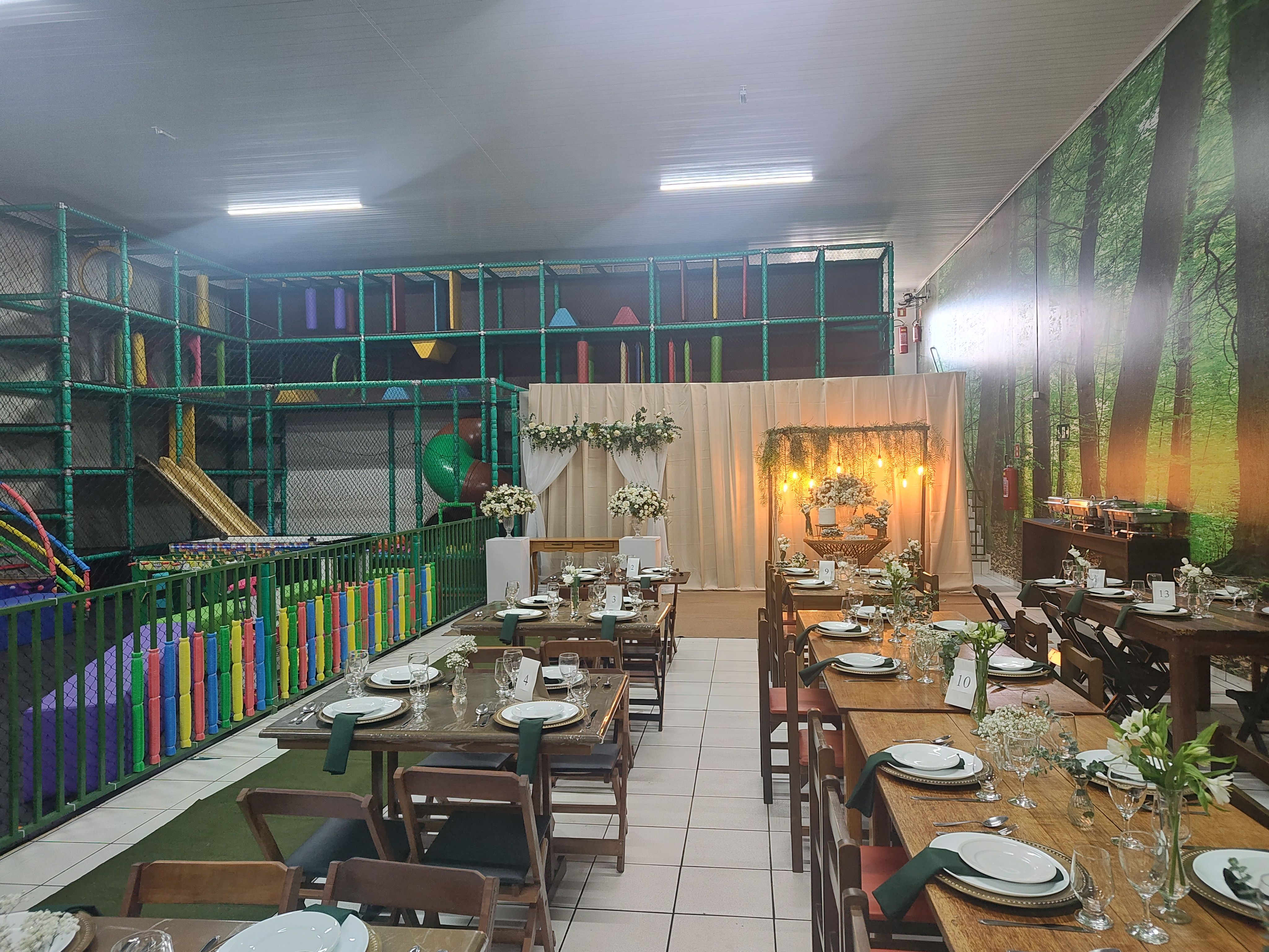 Salão montado para casamento, com mesas de madeira postas, arco de flores ao fundo e o playground à esquerda