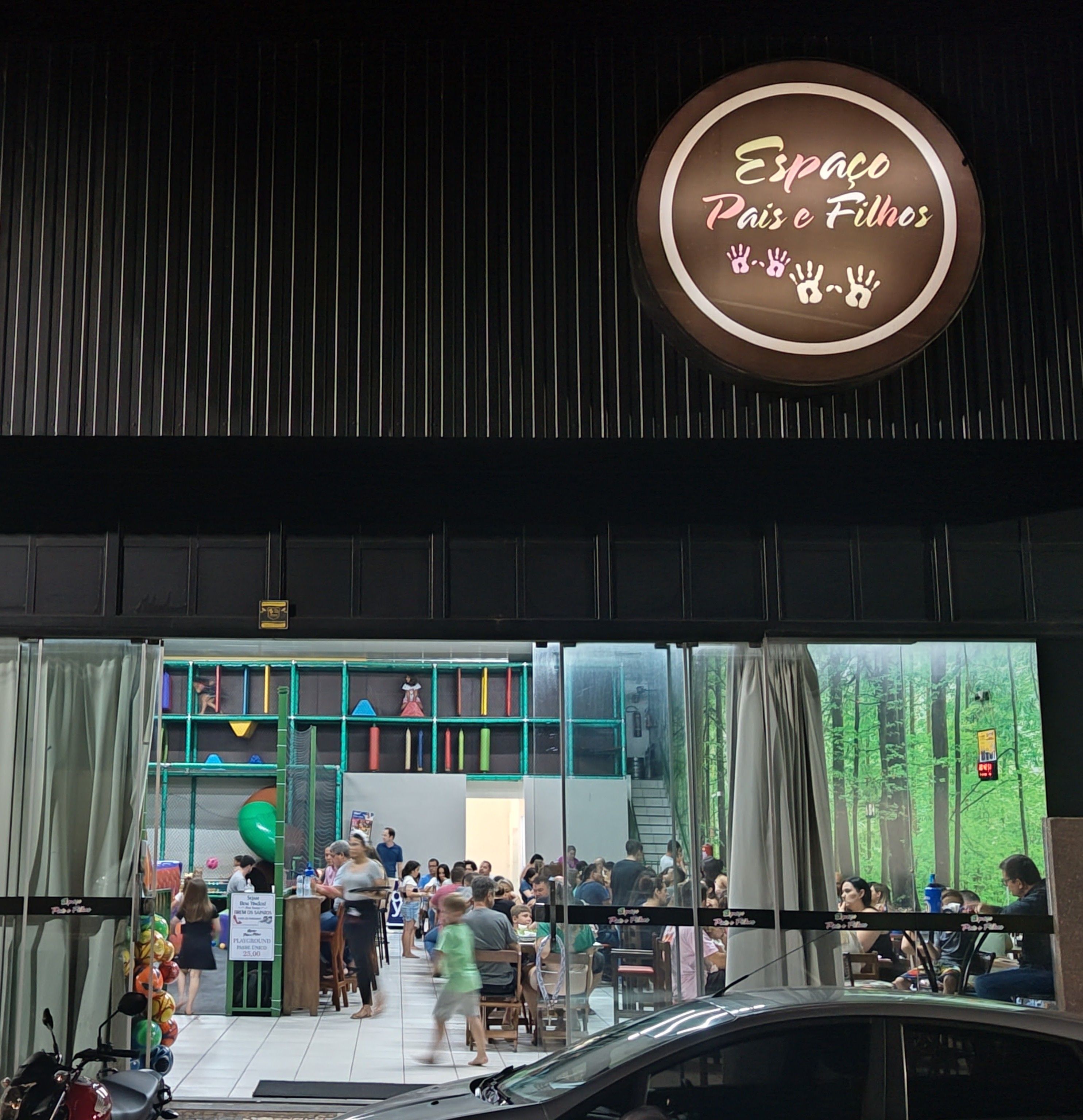 Fachada do Espaço Pais & Filhos à noite, com o letreiro iluminado e o salão cheio visto pela vitrine